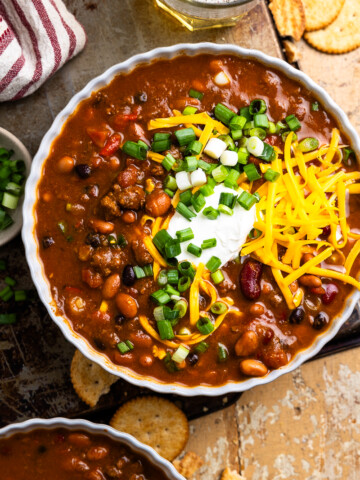 bowl of venison chili topped with cheese and green onions sitting next to a cup of beer and crackers