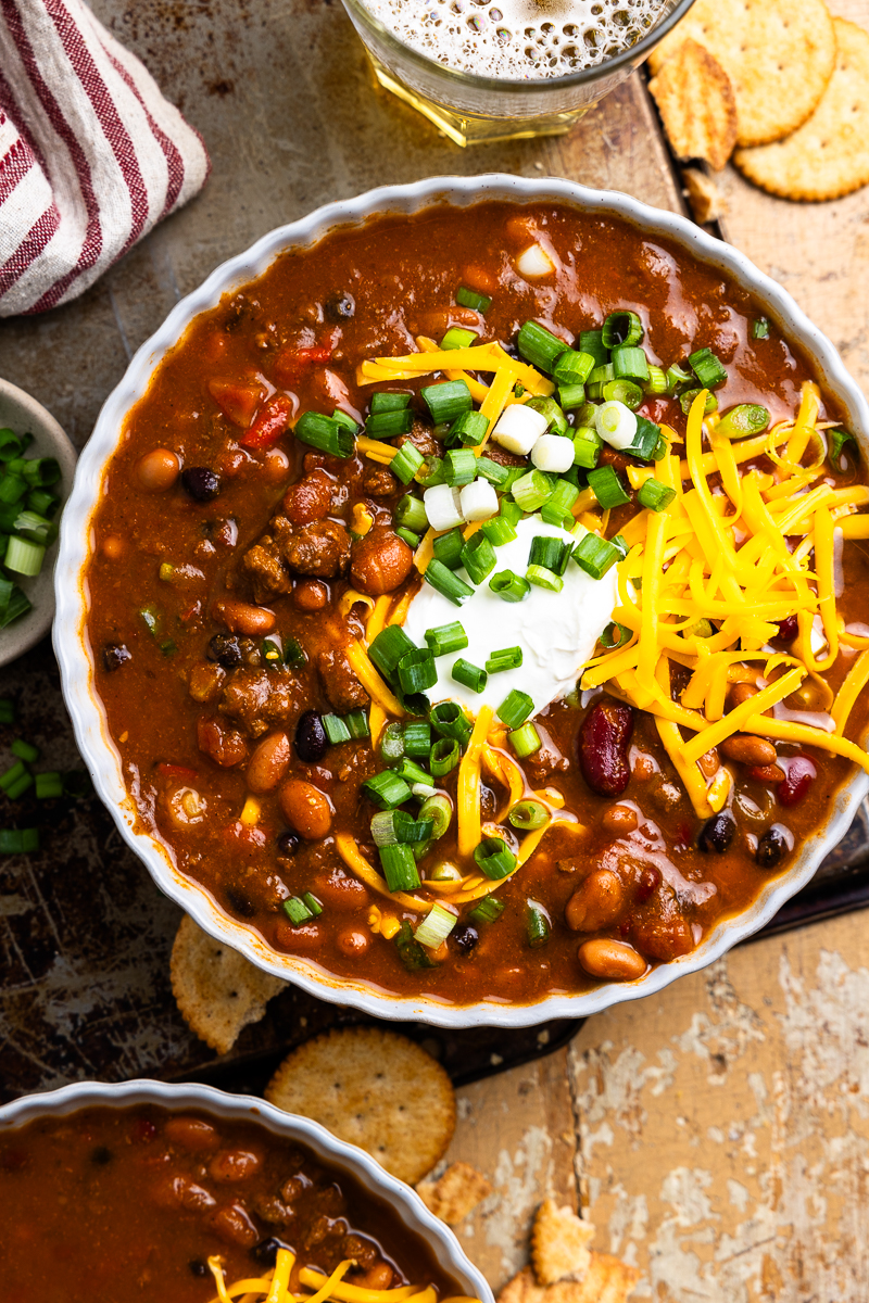 bowl of venison chili topped with cheese and green onions sitting next to a cup of beer and crackers